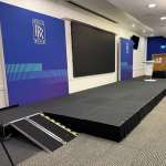 Conference stage with blue Rolls-Royce banners, a large black screen, a podium, and a portable ramp at the front.