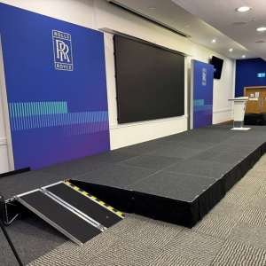 Conference stage with blue Rolls-Royce banners, a large black screen, a podium, and a portable ramp at the front.