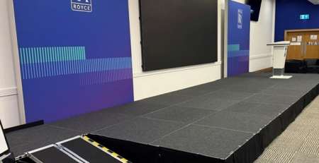 Conference stage with blue Rolls-Royce banners, a large black screen, a podium, and a portable ramp at the front.