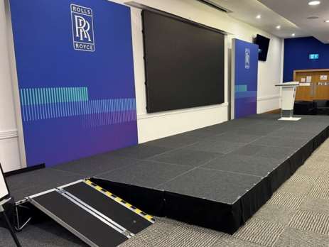 Stage skirting and drapes can improve the appearance of a performance area, hide equipment and create a more professional finish. But in UK venues, the fire performance of these textiles also needs careful consideration. Curtains, drapes, scenery and other combustible materials can contribute to fire risk. GOV.UK fire-safety guidance for places of assembly recommends using Conference stage with blue Rolls-Royce banners, a large black screen, a podium, and a portable ramp at the front.