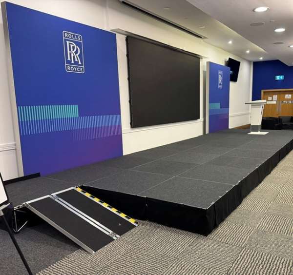 Conference stage with blue Rolls-Royce banners, a large black screen, a podium, and a portable ramp at the front.