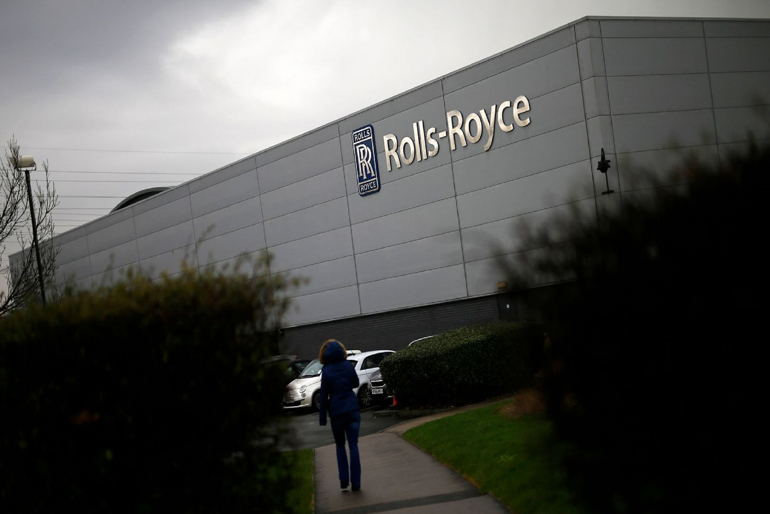 Exterior of a Rolls-Royce facility with the logo on the wall; a person walks along a path in the foreground on a cloudy day.