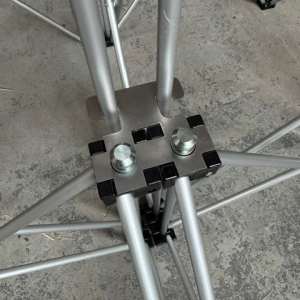 Metal tripod joint with a square metal plate and bolts connecting aluminum tubes on a concrete floor.
