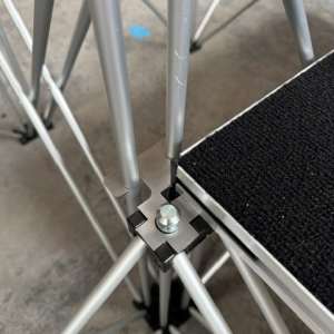 Close-up of a metal scaffolding junction showing a black plastic connector with a bolt securing aluminum tubes and cross-bracing near a textured platform edge.