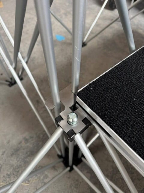 NexGen Riser Leg Link Clamp (Dual) 6 Pack This spare parts connects multi riser legs together of different heights, for use with differing height riser legs where the deck platforms cannot be aligned. Such as a lower step or tier setup. Place the link clamp around the two taller of the two riser legs, and Close-up of a metal scaffolding junction showing a black plastic connector with a bolt securing aluminum tubes and cross-bracing near a textured platform edge.