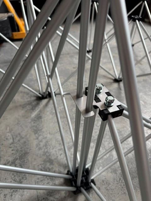 NexGen Riser Leg Link Clamp (Dual) 6 Pack This spare parts connects multi riser legs together of different heights, for use with differing height riser legs where the deck platforms cannot be aligned. Such as a lower step or tier setup. Place the link clamp around the two taller of the two riser legs, and Close-up of gray metal tubing joined by a small black connector plate with two bolts, forming a tubular frame on a concrete floor.
