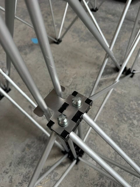 NexGen Riser Leg Link Clamp (Dual) 6 Pack This spare parts connects multi riser legs together of different heights, for use with differing height riser legs where the deck platforms cannot be aligned. Such as a lower step or tier setup. Place the link clamp around the two taller of the two riser legs, and Close-up of a metal scaffold truss joint with a square plate secured by bolts, connecting silver tubes.