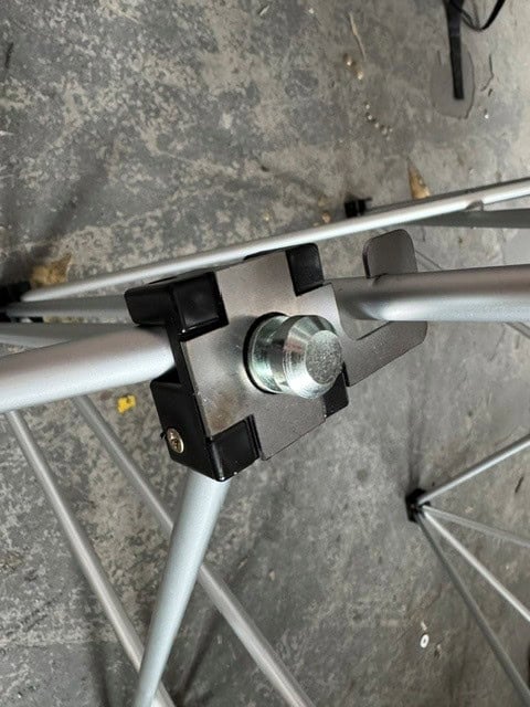 NexGen Riser Leg Link Clamp (Single) 6 Pack This spare parts connects riser legs together of different heights, for use with differing height riser legs where the deck platforms cannot be aligned. Such as a lower step or tier setup. Place the link clamp around the taller of the two riser legs, and over the Close-up of a metal joint with a large bolt and black plastic spacers on a gray folding frame, on a concrete floor.
