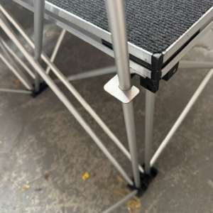 Close-up of a gray metal scaffold platform with a textured black non-slip surface and crossed brace legs on a concrete floor.