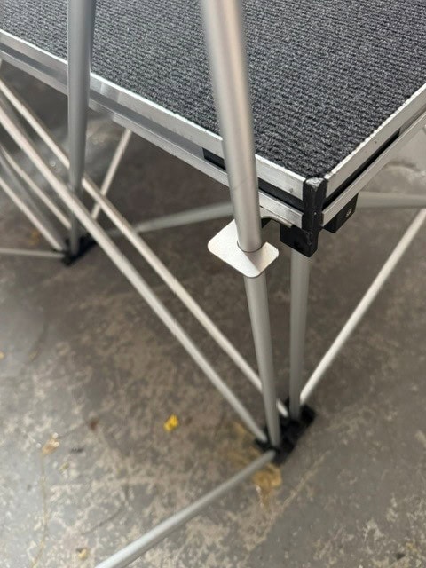 NexGen Riser Leg Link Clamp (Single) 6 Pack This spare parts connects riser legs together of different heights, for use with differing height riser legs where the deck platforms cannot be aligned. Such as a lower step or tier setup. Place the link clamp around the taller of the two riser legs, and over the Close-up of a gray metal scaffold platform with a textured black non-slip surface and crossed brace legs on a concrete floor.