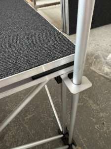 NexGen Riser Leg Link Clamp (Single) 6 Pack This spare parts connects riser legs together of different heights, for use with differing height riser legs where the deck platforms cannot be aligned. Such as a lower step or tier setup. Place the link clamp around the taller of the two riser legs, and over the Close-up of a portable stage edge with a carpeted deck, metal frame, and a locking leg clamp in place.
