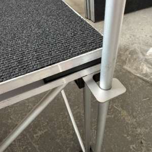 Close-up of a portable stage edge with a carpeted deck, metal frame, and a locking leg clamp in place.