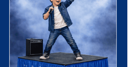 Young boy performing on a stage with a microphone, wearing sunglasses and a denim jacket, raising one arm in the spotlight.