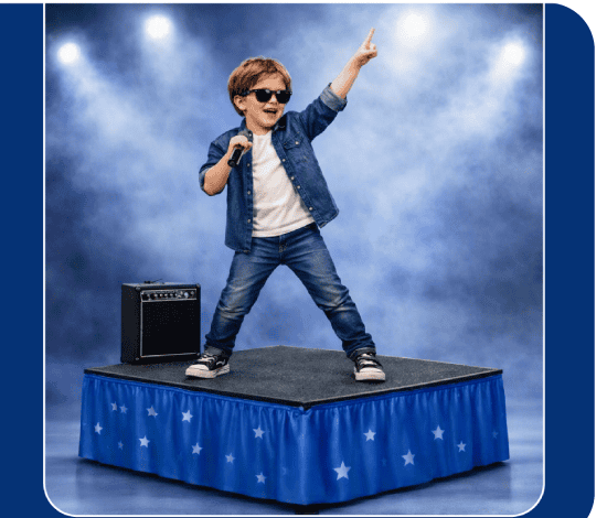 Young boy performing on a stage with a microphone, wearing sunglasses and a denim jacket, raising one arm in the spotlight.
